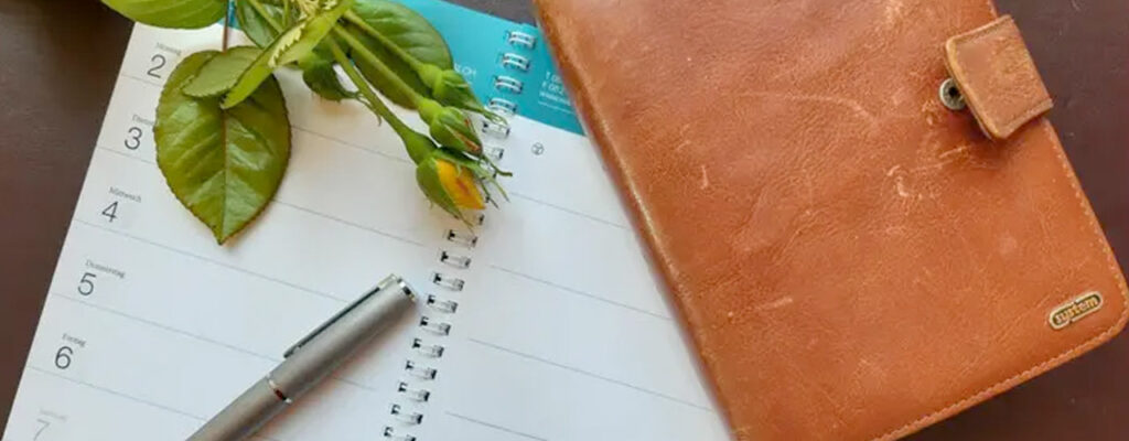 aufgeschlagener Terminkalender und geschlossener Terminplaner mit Rosenknospen und silbernem Stift
