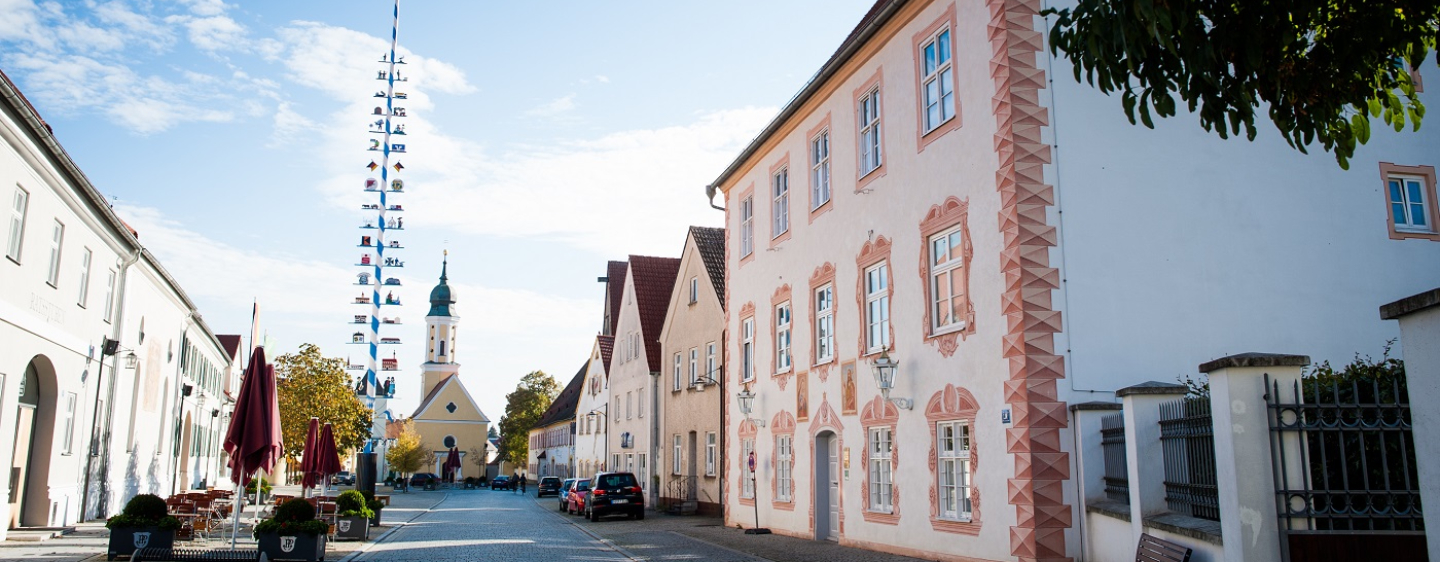 Marktplatz in Pöttmes mit kunstvoll bemaltem Gebäude, Maibaum, Kirche und Biergarten