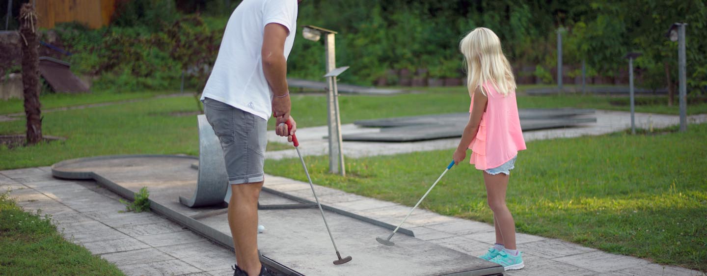 Vater spielt mit Tochter Minigolf, beide halten einen Schläger in den Händen und blicken in Ballrichtung auf das Hindernis. Im Hintergrund sind weitere Spielstationen der Minigolfanlage zu sehen.