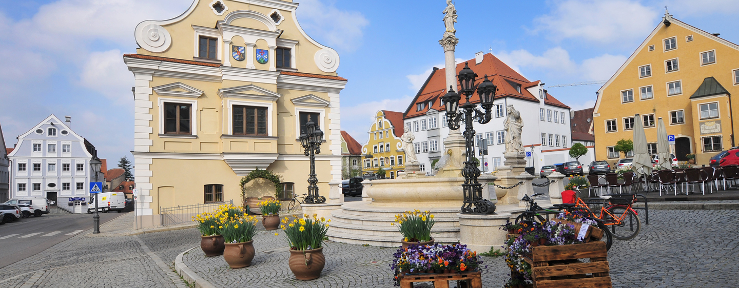 Friedberger Stadtplazt. Im Hintergrund befinden sich historische Gebäude. In der linken Bildhälfte das mit Verschnörkelungen dekorierte, baroke Rathaus. Im Vordergrund ist ein Brunnen, umgeben von Blumendekoration.