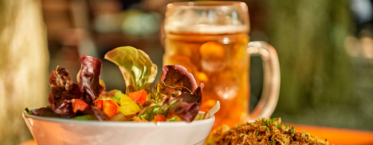 Im Vordergrund ist ein Salat in einer Schüssel angerichtet, daneben steht ein Teller mit Röstzwiebeln. Im Hintergrund eine volle Maß Bier.