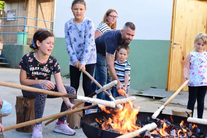 Kinder grillen Stockbrot am Lagerfeuer auf dem Erlebnisbauernhof beim Hibsch. Die Kinder halten Stöcke mit Brot ins Feuer einer Feuerschale, die in der Mitte steht.