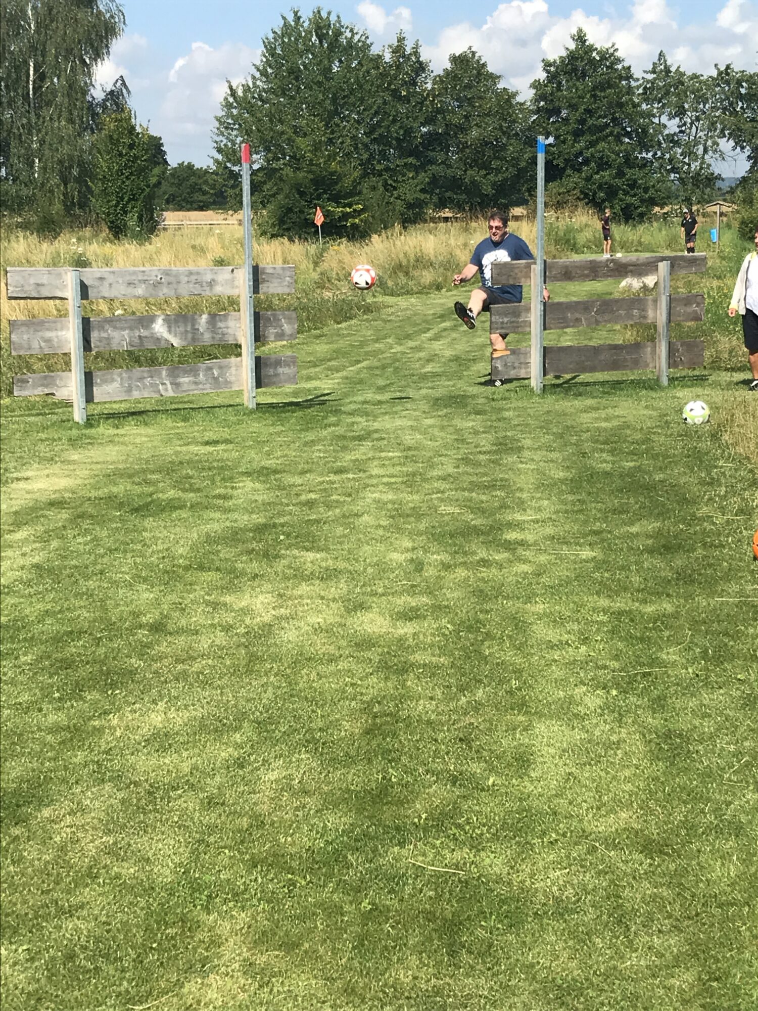 Mann kickt Fußball durch einen Zaun auf einer Wiese des Soccerparks Rehling, im Hintergrund weitere Spieler