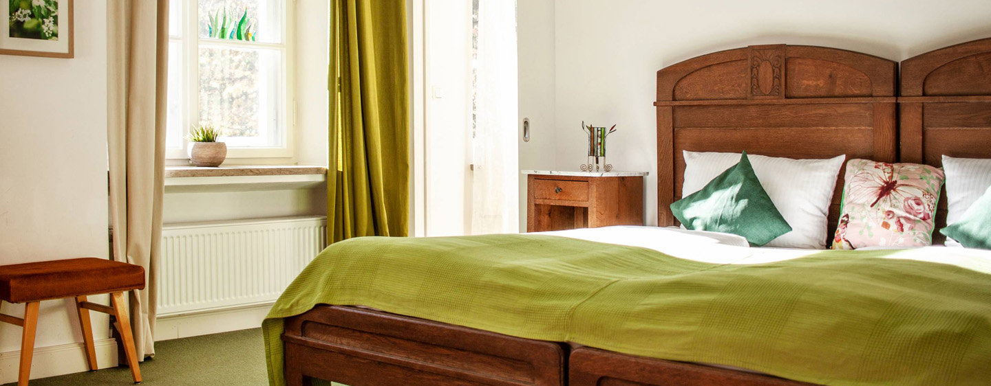 Innenansicht Hotelzimmer. Holzbett mit grüner Decke, grünen Kissen. Grüne Vorhänege.