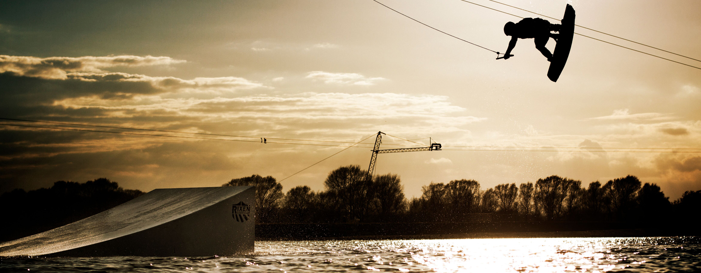 Kitboarder springt bei Sonnenuntergang über eine Rampe auf der Wasserskianlage Chill & Wake
