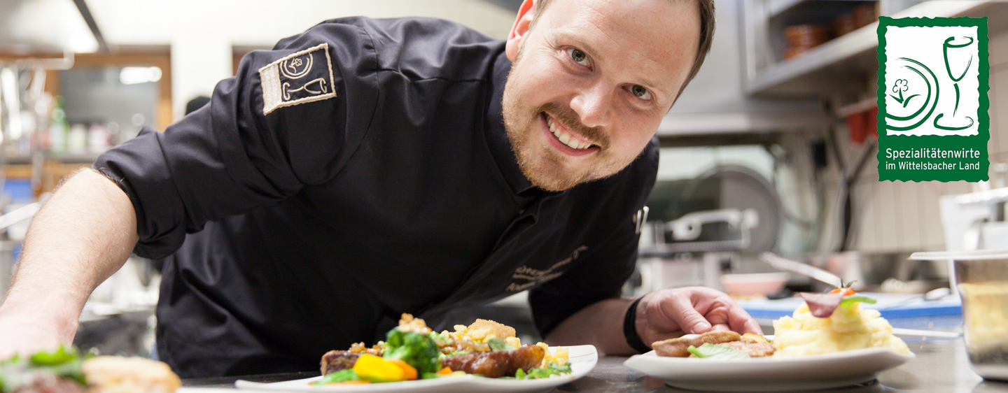 Koch blickt durch die Anrichte mit zwei fertig gedeckten Tellern. Am Bildrand das Logo der "Spezialitätenwirte im Wittelsbacher Land"