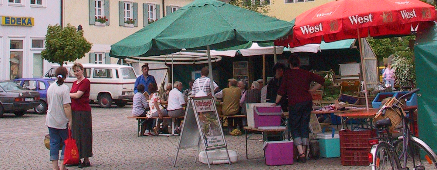 Wochenmarkt in Kühbach. Verschiedene Stände sind aufgebaut. An einem Stand sitzen einige Besucher auf Bierbänken. Im Hintergrund Parkplätze und ein Supermarkt.