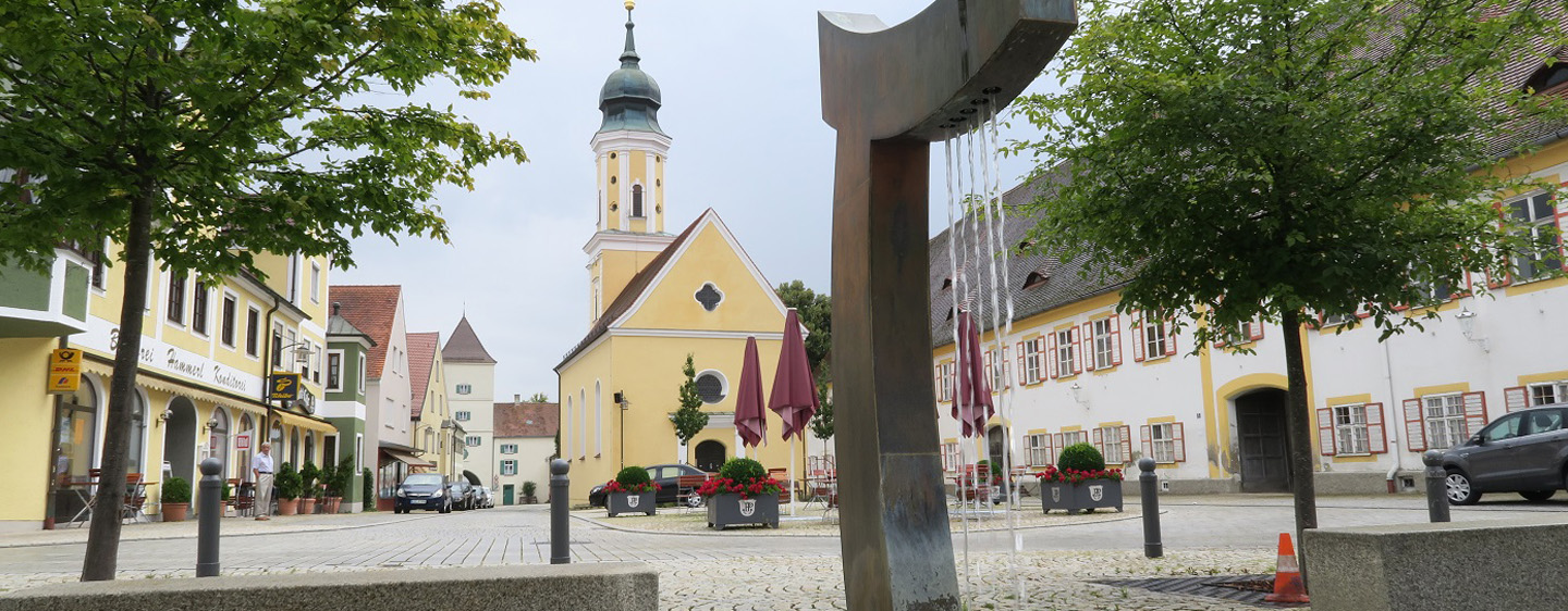 Marktplatz in Pöttmes. Historische Altstadt mit Blick auf eine Kirche. Davor Tische, Stühle und geschlossene Schirm eines Gasthofs
