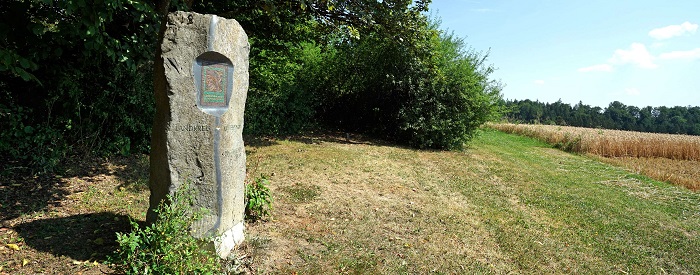 Gedenkstein Mittelpunkt des Landkreises Aichach-Friedberg auf eine Wiese mit Hecke daneben ein Feld