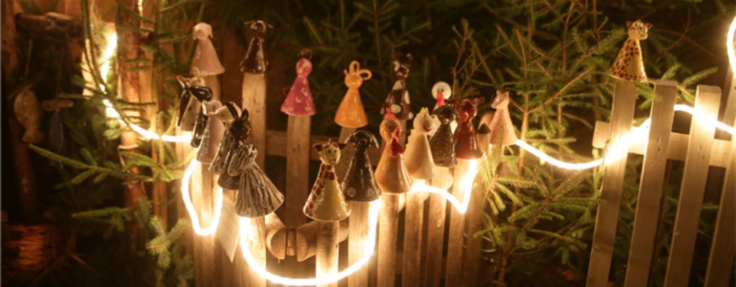 getöpferte Figuren stehen auf einem Holzzaun der weihnachtlich mit Tannenzweitgen dekoriert und beleuchtet ist.