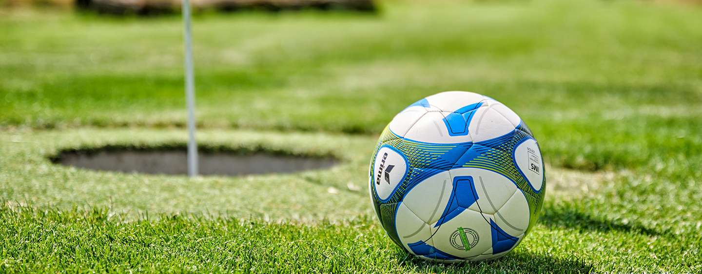 Ein Fußball liegt auf einer Wiese vor einem übergroßen Golf-Loch. Die Sportart nennt sich Soccergolf.