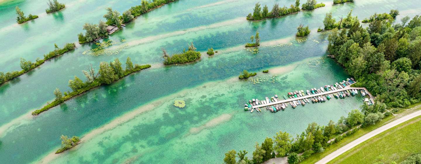Luftaufnahme des Weitmannsees. Im See liegen viele kleine Inseln und nahe des Ufers schwimmt ein langer Steg an dem viele Fischerboote anliegen. Das Wasser ist türkis bis smaragdgrün.