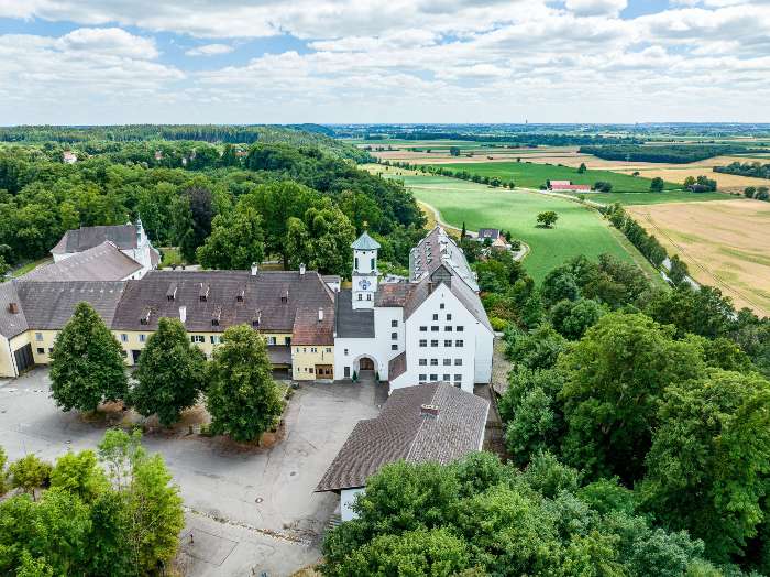 Luftaufnahme des Schlosses Schnerneck in Rehling. Großer weißer gebäudekomplex mit vielen Fenstern. Dahinter erstreckt sich die Lechebene.