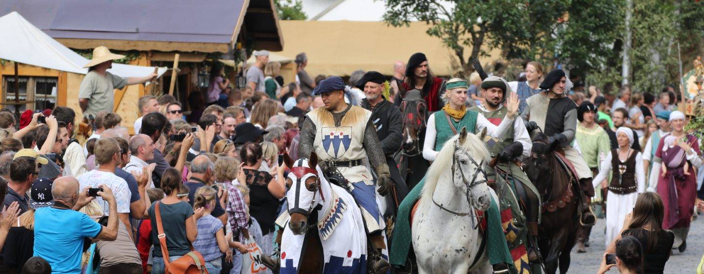 Menschen in historischen Gewänden reiten auf Pferden bei einem Umzug umringt von Zuschauern
