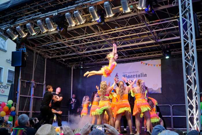 Eine Gruppe kostümierter Frauen, die auf einer Bühne stehen, fangen eine durch die Luft fliegende kostümierte Frau auf.